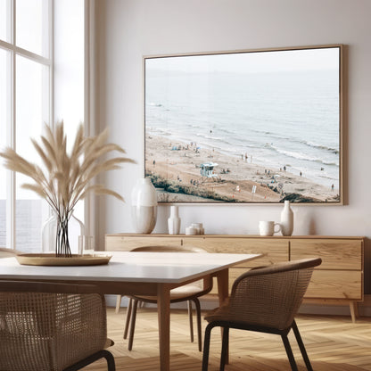 A slightly hazy, high-angle Landscape Beach Print captures a wide beachscape print where a light blue ocean waves meet a sandy shore dotted with numerous people enjoying the Summer Vibes. Gentle white waves roll towards the beach. A Blue Lifeguard Tower stands prominently in the lower center of the framed landscape beach print. The overall lighting suggests a bright, overcast travel photography day on the San Diego coast. Photograph in minimalist dining room mock up