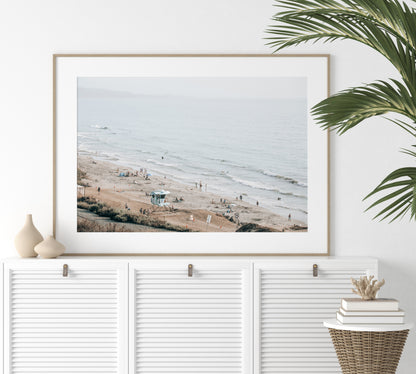 A slightly hazy, high-angle Landscape Beach Print captures a wide beachscape print where a light blue ocean waves meet a sandy shore dotted with numerous people enjoying the Summer Vibes. Gentle white waves roll towards the beach. A Blue Lifeguard Tower stands prominently in the lower center of the framed landscape beach print. The overall lighting suggests a bright, overcast travel photography day on the San Diego coast. Photograph in minimalist mock up