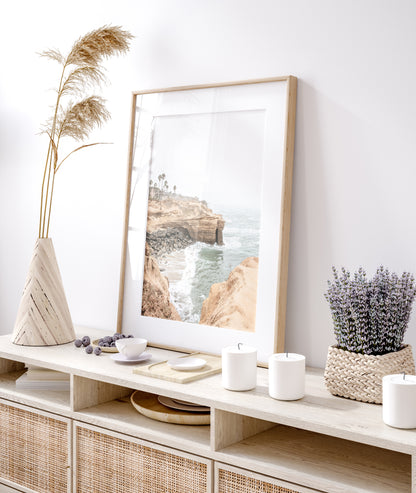 Fine art of Sunset Cliffs, San Diego California vertical print of hazy high-angle landscape beach photography captures a light blue ocean meeting tall tan, rocky, brown cliffs rise as iconic coastal scenery. Photograph in minimalist mock up.
