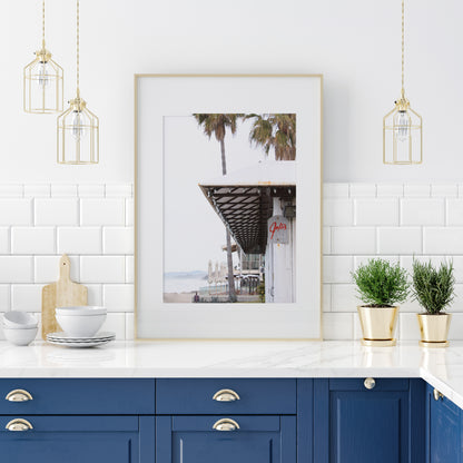 A framed beach scene art print with a wooden structure and palm trees, titled 'Jake's' on the sign, set against a background of a beach and ocean. Photograph in minimalist mock up.