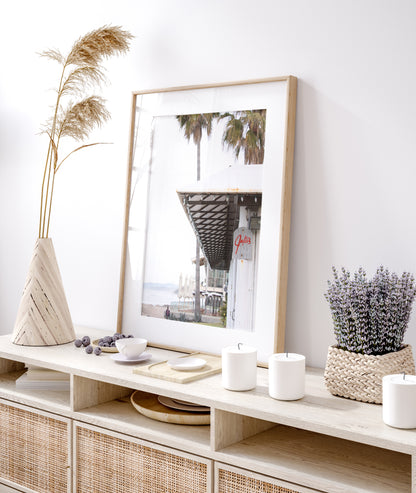 A framed beach scene art print with a wooden structure and palm trees, titled 'Jake's' on the sign, set against a background of a beach and ocean. Photograph in minimalist mock up.
