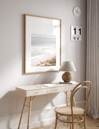 Minimalist ocean print of ocean waves with surfers in the distance and a sandy hillside in the foreground. 