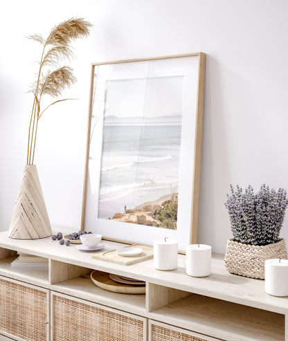 Minimalist ocean print of ocean waves with surfers in the distance and a sandy hillside in the foreground. 
