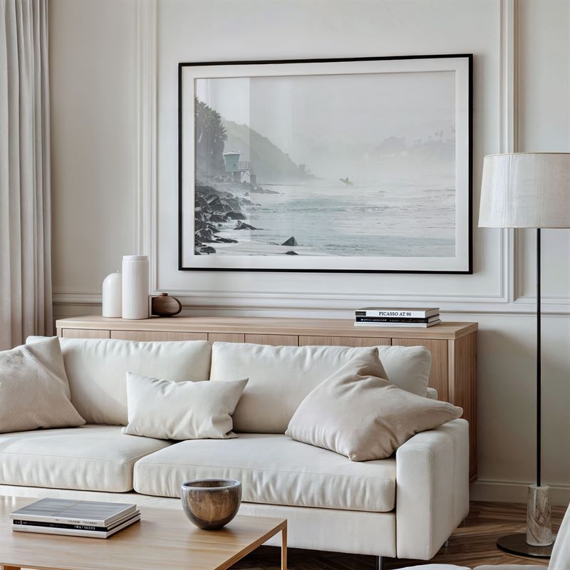 Modern living room with a white sofa, wooden coffee table, and framed artwork on the wall of a coastal scene.