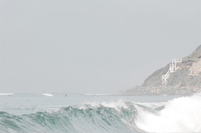 Neutral ocean print featuring a beautifully captured turquoise wave cresting with white seafoam, embodying minimalist surf art. This horizontal coastal photography showcases soft, pastel tones under a hazy sky. In the background, a rugged cliffside staircase on the right meets the sea, with faint distant surfers in the water, suggesting the expansive Southern California coastline. A tranquil piece of surf wall art, perfect for a coastal theme.