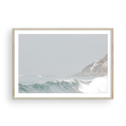 Neutral ocean print featuring a beautifully captured turquoise wave cresting with white seafoam, embodying minimalist surf art. This horizontal coastal photography showcases soft, pastel tones under a hazy sky. In the background, a rugged cliffside staircase on the right meets the sea, with faint distant surfers in the water, suggesting the expansive Southern California coastline. A tranquil piece of surf wall art, perfect for a coastal theme. Photograph in framed minimalist mock up.