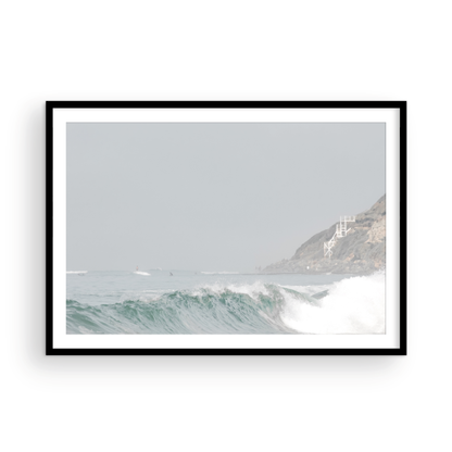 Neutral ocean print featuring a beautifully captured turquoise wave cresting with white seafoam, embodying minimalist surf art. This horizontal coastal photography showcases soft, pastel tones under a hazy sky. In the background, a rugged cliffside staircase on the right meets the sea, with faint distant surfers in the water, suggesting the expansive Southern California coastline. A tranquil piece of surf wall art, perfect for a coastal theme. Photograph in framed minimalist mock up.