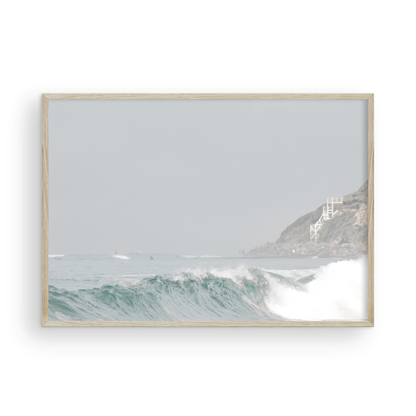 Neutral ocean print featuring a beautifully captured turquoise wave cresting with white seafoam, embodying minimalist surf art. This horizontal coastal photography showcases soft, pastel tones under a hazy sky. In the background, a rugged cliffside staircase on the right meets the sea, with faint distant surfers in the water, suggesting the expansive Southern California coastline. A tranquil piece of surf wall art, perfect for a coastal theme. Photograph in framed minimalist mock up.