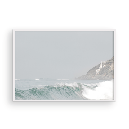 Neutral ocean print featuring a beautifully captured turquoise wave cresting with white seafoam, embodying minimalist surf art. This horizontal coastal photography showcases soft, pastel tones under a hazy sky. In the background, a rugged cliffside staircase on the right meets the sea, with faint distant surfers in the water, suggesting the expansive Southern California coastline. A tranquil piece of surf wall art, perfect for a coastal theme. Photograph in framed minimalist mock up.