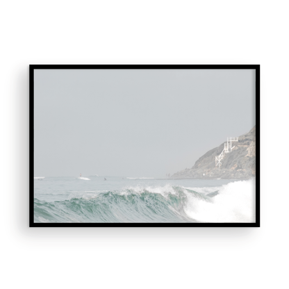 Neutral ocean print featuring a beautifully captured turquoise wave cresting with white seafoam, embodying minimalist surf art. This horizontal coastal photography showcases soft, pastel tones under a hazy sky. In the background, a rugged cliffside staircase on the right meets the sea, with faint distant surfers in the water, suggesting the expansive Southern California coastline. A tranquil piece of surf wall art, perfect for a coastal theme. Photograph in framed minimalist mock up.