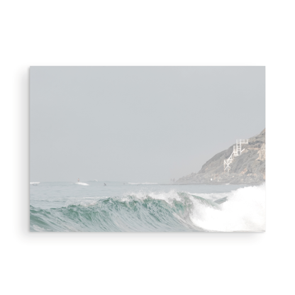 Neutral ocean print featuring a beautifully captured turquoise wave cresting with white seafoam, embodying minimalist surf art. This horizontal coastal photography showcases soft, pastel tones under a hazy sky. In the background, a rugged cliffside staircase on the right meets the sea, with faint distant surfers in the water, suggesting the expansive Southern California coastline. A tranquil piece of surf wall art, perfect for a coastal theme. Photograph in unframed minimalist mock up.