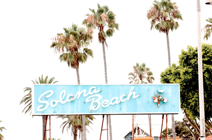 Colorful travel photography capturing the iconic 'Solana Beach' sign, a quintessential piece of Surf Signs San Diego art. The retro light blue sign, adorned with white cursive script and a unique steer head logo, is set against a backdrop of towering palm trees and a bright, hazy California sky. This Southern California surf beach sign evokes strong summer vibes and is perfect for coastal decor. 