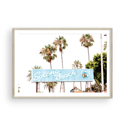 Colorful travel photography capturing the iconic 'Solana Beach' sign, a quintessential piece of Surf Signs San Diego art. The retro light blue sign, adorned with white cursive script and a unique steer head logo, is set against a backdrop of towering palm trees and a bright, hazy California sky. This Southern California surf beach sign evokes strong summer vibes and is perfect for coastal decor. Photograph in minimalist framed mock up.
