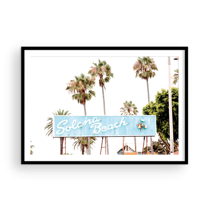 Colorful travel photography capturing the iconic 'Solana Beach' sign, a quintessential piece of Surf Signs San Diego art. The retro light blue sign, adorned with white cursive script and a unique steer head logo, is set against a backdrop of towering palm trees and a bright, hazy California sky. This Southern California surf beach sign evokes strong summer vibes and is perfect for coastal decor. Photograph in minimalist framed mock up.