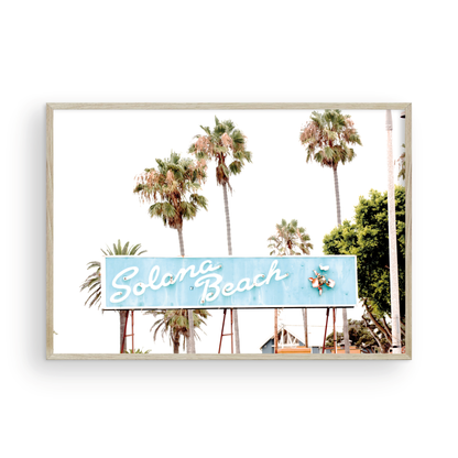 Colorful travel photography capturing the iconic 'Solana Beach' sign, a quintessential piece of Surf Signs San Diego art. The retro light blue sign, adorned with white cursive script and a unique steer head logo, is set against a backdrop of towering palm trees and a bright, hazy California sky. This Southern California surf beach sign evokes strong summer vibes and is perfect for coastal decor. Photograph in minimalist framed mock up.