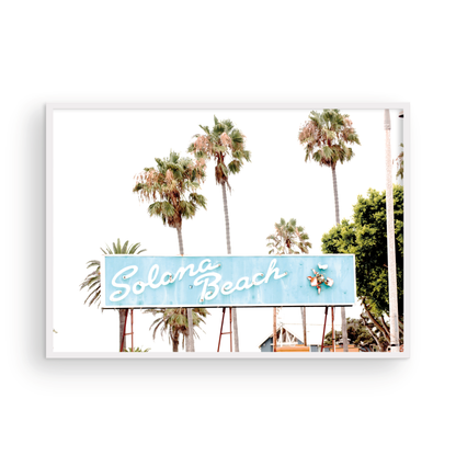 Colorful travel photography capturing the iconic 'Solana Beach' sign, a quintessential piece of Surf Signs San Diego art. The retro light blue sign, adorned with white cursive script and a unique steer head logo, is set against a backdrop of towering palm trees and a bright, hazy California sky. This Southern California surf beach sign evokes strong summer vibes and is perfect for coastal decor. Photograph in minimalist framed mock up.
