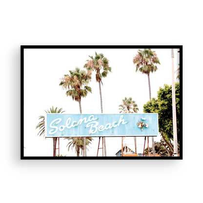 Colorful travel photography capturing the iconic 'Solana Beach' sign, a quintessential piece of Surf Signs San Diego art. The retro light blue sign, adorned with white cursive script and a unique steer head logo, is set against a backdrop of towering palm trees and a bright, hazy California sky. This Southern California surf beach sign evokes strong summer vibes and is perfect for coastal decor. Photograph in minimalist framed mock up.