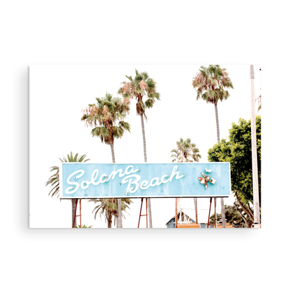 Colorful travel photography capturing the iconic 'Solana Beach' sign, a quintessential piece of Surf Signs San Diego art. The retro light blue sign, adorned with white cursive script and a unique steer head logo, is set against a backdrop of towering palm trees and a bright, hazy California sky. This Southern California surf beach sign evokes strong summer vibes and is perfect for coastal decor. Photograph in minimalist framed mock up.