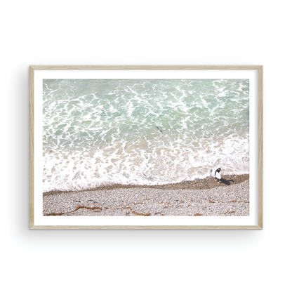 Horizontal surf print of a surfer with a surfboard walking along a rocky beach next to foamy ocean waves. This surfing art captures a unique coastline. Photograph in minimalist mock up.