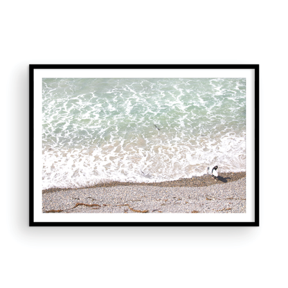 Horizontal surf print of a surfer with a surfboard walking along a rocky beach next to foamy ocean waves. This surfing art captures a unique coastline. Photograph in minimalist mock up.