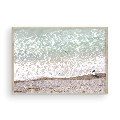 Horizontal surf print of a surfer with a surfboard walking along a rocky beach next to foamy ocean waves. This surfing art captures a unique coastline. Photograph in minimalist mock up.