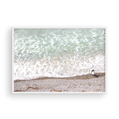 Horizontal surf print of a surfer with a surfboard walking along a rocky beach next to foamy ocean waves. This surfing art captures a unique coastline. Photograph in minimalist mock up.