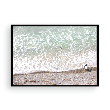 Horizontal surf print of a surfer with a surfboard walking along a rocky beach next to foamy ocean waves. This surfing art captures a unique coastline. Photograph in minimalist mock up.