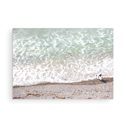 Horizontal surf print of a surfer with a surfboard walking along a rocky beach next to foamy ocean waves. This surfing art captures a unique coastline. Photograph in minimalist mock up.