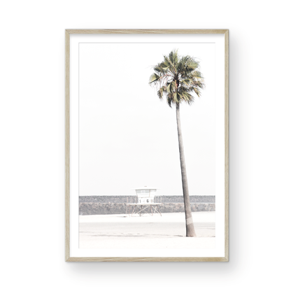 Dreamy coastal palm tree print featuring a tall palm tree in the foreground and a pastel blue lifeguard tower centered in the distance. This vertical tropical print is captured at Oceanside Beach on the Southern California coast, is bathed in bright, hazy sunlight, creating a pastel beach art effect. Photograph in minimalist mock up.