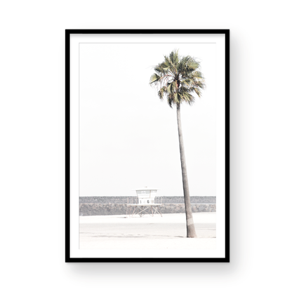 Dreamy coastal palm tree print featuring a tall palm tree in the foreground and a pastel blue lifeguard tower centered in the distance. This vertical tropical print is captured at Oceanside Beach on the Southern California coast, is bathed in bright, hazy sunlight, creating a pastel beach art effect. Photograph in minimalist mock up.
