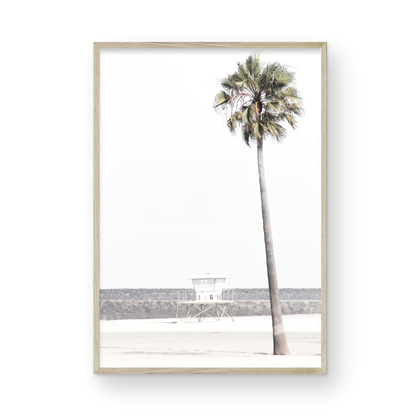 Dreamy coastal palm tree print featuring a tall palm tree in the foreground and a pastel blue lifeguard tower centered in the distance. This vertical tropical print is captured at Oceanside Beach on the Southern California coast, is bathed in bright, hazy sunlight, creating a pastel beach art effect. Photograph in minimalist mock up.