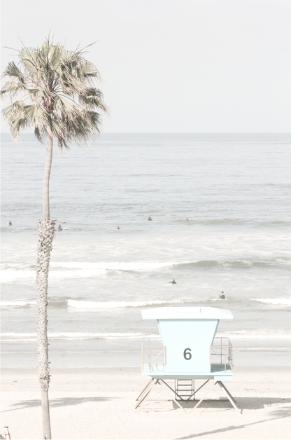 Dreamy coastal palm tree print featuring a tall palm tree with shaggy trunks in the foreground and a pastel blue lifeguard tower centered in the distance. This vertical tropical print is captured at Oceanside Beach on the Southern California coast, is bathed in bright, hazy sunlight, creating a pastel beach art effect.