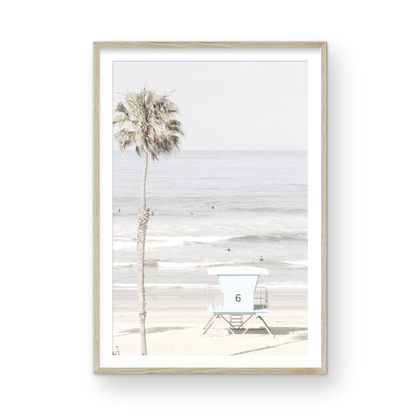 Dreamy coastal palm tree print featuring a tall palm tree with shaggy trunks in the foreground and a pastel blue lifeguard tower centered in the distance. This vertical tropical print is captured at Oceanside Beach on the Southern California coast, is bathed in bright, hazy sunlight, creating a pastel beach art effect. Photograph in minimalist mock up.