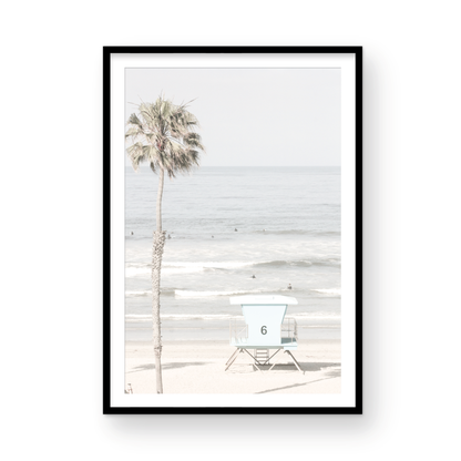 Dreamy coastal palm tree print featuring a tall palm tree with shaggy trunks in the foreground and a pastel blue lifeguard tower centered in the distance. This vertical tropical print is captured at Oceanside Beach on the Southern California coast, is bathed in bright, hazy sunlight, creating a pastel beach art effect. Photograph in minimalist mock up.