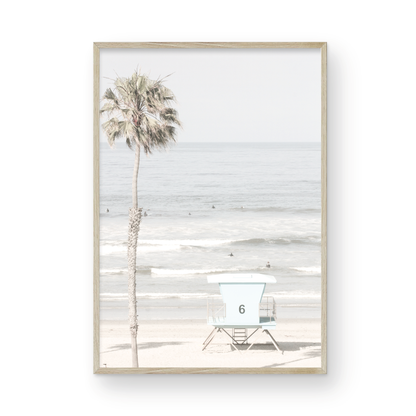 Dreamy coastal palm tree print featuring a tall palm tree with shaggy trunks in the foreground and a pastel blue lifeguard tower centered in the distance. This vertical tropical print is captured at Oceanside Beach on the Southern California coast, is bathed in bright, hazy sunlight, creating a pastel beach art effect. Photograph in minimalist mock up.