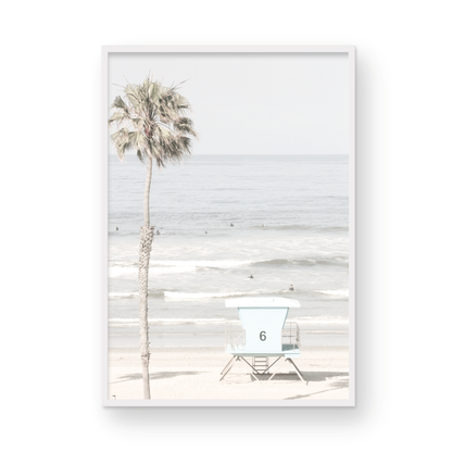 Dreamy coastal palm tree print featuring a tall palm tree with shaggy trunks in the foreground and a pastel blue lifeguard tower centered in the distance. This vertical tropical print is captured at Oceanside Beach on the Southern California coast, is bathed in bright, hazy sunlight, creating a pastel beach art effect. Photograph in minimalist mock up.