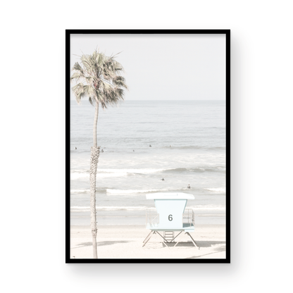Dreamy coastal palm tree print featuring a tall palm tree with shaggy trunks in the foreground and a pastel blue lifeguard tower centered in the distance. This vertical tropical print is captured at Oceanside Beach on the Southern California coast, is bathed in bright, hazy sunlight, creating a pastel beach art effect. Photograph in minimalist mock up.
