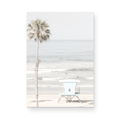 Dreamy coastal palm tree print featuring a tall palm tree with shaggy trunks in the foreground and a pastel blue lifeguard tower centered in the distance. This vertical tropical print is captured at Oceanside Beach on the Southern California coast, is bathed in bright, hazy sunlight, creating a pastel beach art effect. Photograph in minimalist mock up.