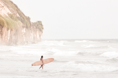 Horizontal surf art print of a lone surfer carrying a surfboard, walking on a rocky, foggy shoreline at Moonlight State Beach with a cliff in the background. 