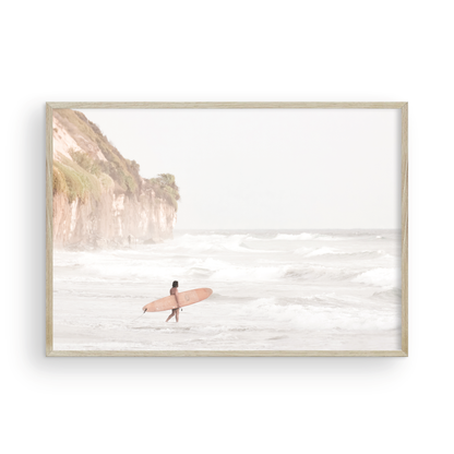 Horizontal surf art print of a lone surfer carrying a surfboard, walking on a rocky, foggy shoreline at Moonlight State Beach with a cliff in the background. Photograph in minimalist mock up.