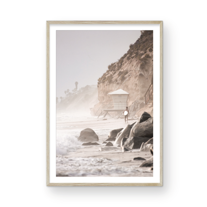 Vertical surf art print of a lone surfer carrying a surfboard, walking on a rocky, foggy shoreline at Moonlight State Beach with a lifeguard tower and cliff in the background. Photograph in minimalist mock up.