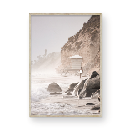 Vertical surf art print of a lone surfer carrying a surfboard, walking on a rocky, foggy shoreline at Moonlight State Beach with a lifeguard tower and cliff in the background. Photograph in minimalist mock up.