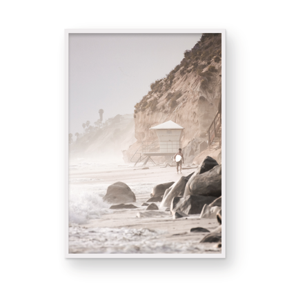 Vertical surf art print of a lone surfer carrying a surfboard, walking on a rocky, foggy shoreline at Moonlight State Beach with a lifeguard tower and cliff in the background. Photograph in minimalist mock up.