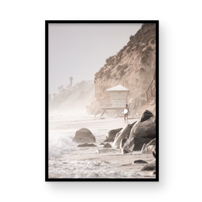 Vertical surf art print of a lone surfer carrying a surfboard, walking on a rocky, foggy shoreline at Moonlight State Beach with a lifeguard tower and cliff in the background. Photograph in minimalist mock up.