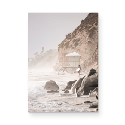 Vertical surf art print of a lone surfer carrying a surfboard, walking on a rocky, foggy shoreline at Moonlight State Beach with a lifeguard tower and cliff in the background. Photograph in minimalist mock up.