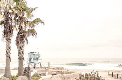 Dreamy coastal palm tree print featuring two tall palm trees with shaggy trunks in the foreground and a pastel blue lifeguard tower in the distance. This horizontal tropical print is captured at Seaside Beach on the Southern California coast, is bathed in bright, hazy sunlight, creating a pastel beach art effect. Evokes serene summer vibes, perfect for beach house decor.
