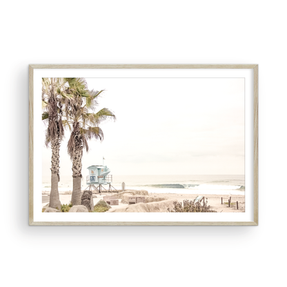 Dreamy coastal palm tree print featuring two tall palm trees with shaggy trunks in the foreground and a pastel blue lifeguard tower in the distance. This horizontal tropical print is captured at Seaside Beach on the Southern California coast, is bathed in bright, hazy sunlight, creating a pastel beach art effect. Evokes serene summer vibes, perfect for beach house decor. Photograph in minimalist mock up.