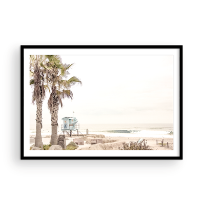 Dreamy coastal palm tree print featuring two tall palm trees with shaggy trunks in the foreground and a pastel blue lifeguard tower in the distance. This horizontal tropical print is captured at Seaside Beach on the Southern California coast, is bathed in bright, hazy sunlight, creating a pastel beach art effect. Evokes serene summer vibes, perfect for beach house decor. Photograph in minimalist mock up.