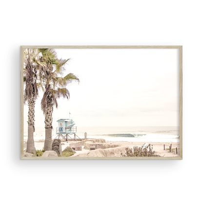 Dreamy coastal palm tree print featuring two tall palm trees with shaggy trunks in the foreground and a pastel blue lifeguard tower in the distance. This horizontal tropical print is captured at Seaside Beach on the Southern California coast, is bathed in bright, hazy sunlight, creating a pastel beach art effect. Evokes serene summer vibes, perfect for beach house decor. Photograph in minimalist mock up.