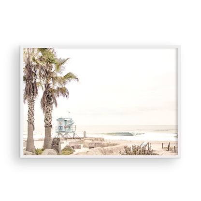 Dreamy coastal palm tree print featuring two tall palm trees with shaggy trunks in the foreground and a pastel blue lifeguard tower in the distance. This horizontal tropical print is captured at Seaside Beach on the Southern California coast, is bathed in bright, hazy sunlight, creating a pastel beach art effect. Evokes serene summer vibes, perfect for beach house decor. Photograph in minimalist mock up.